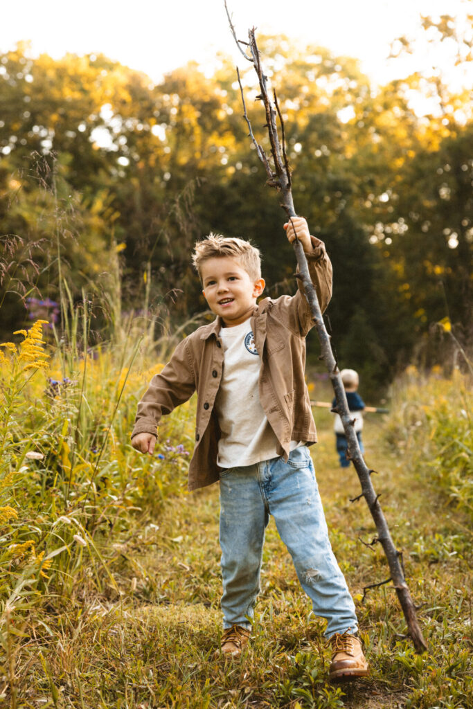 Young boy in a tan jacket and jeans holding up a tall stick in a golden meadow
