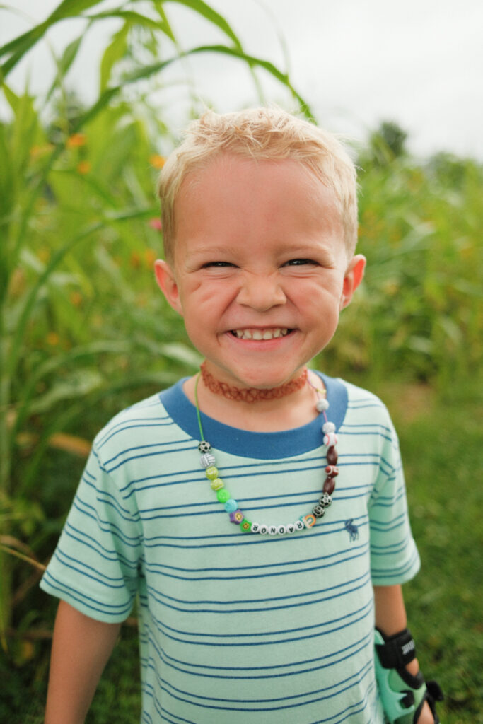 Grinning blond boy in a green striped tee wearing a beaded name necklace by tall corn
