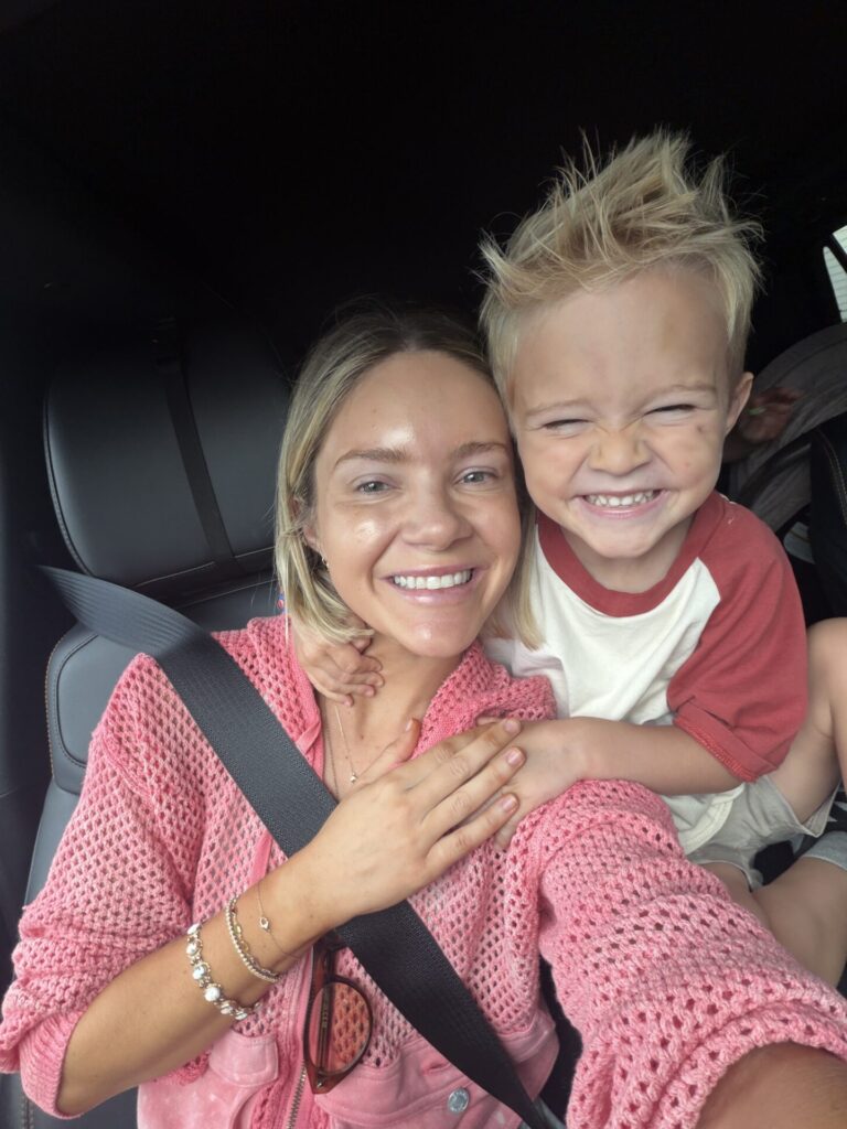 Hunter in a pink knit top taking a car selfie with a laughing blond boy in red