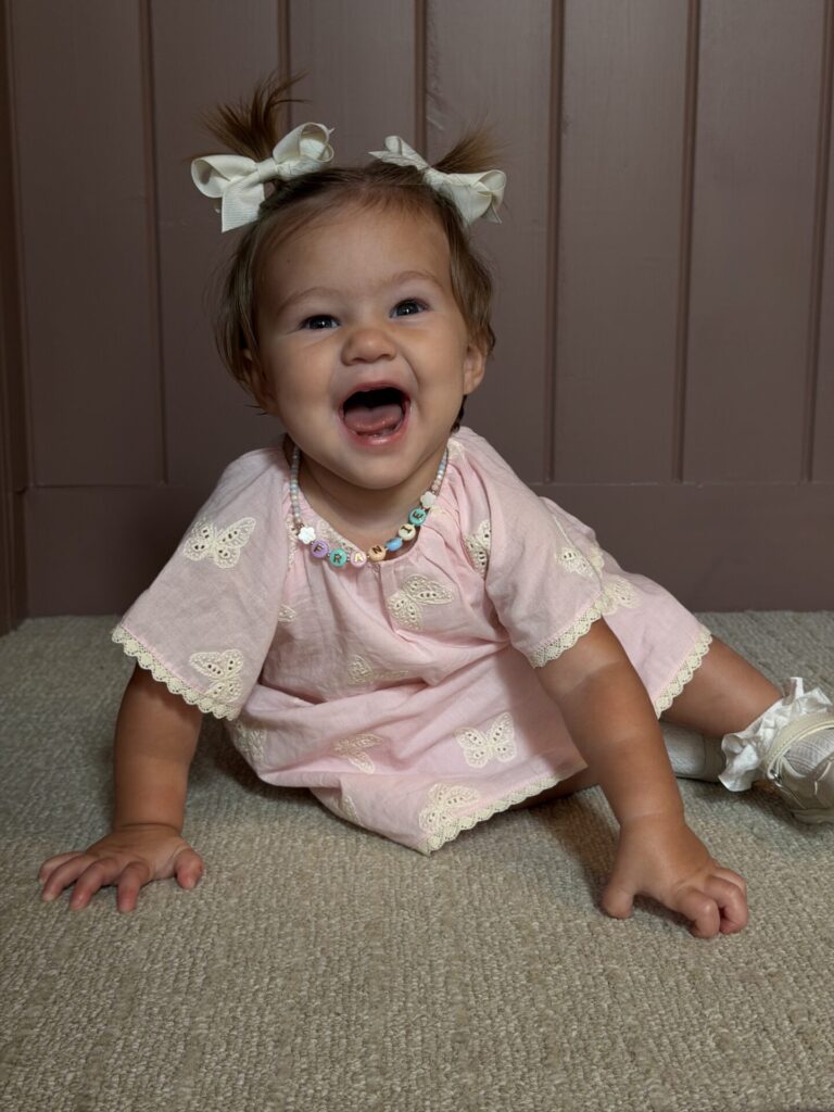 Laughing baby girl in a pink butterfly dress and cream pigtail bows sitting on carpet